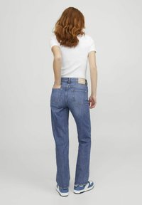 Mėlyni džinsiniai platėjantys džinsai su klasikos penkių kišenių dizainu ir siuvinėjimu, derinami su balta prigludusia marškinėlių ir sportinėmis bateliais.