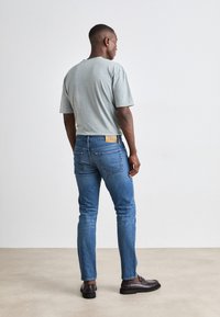 Siva pamučna majica, kratki rukavi, u kombinaciji s plavim trapericama, slim fit, i smešnim kožnim cipelama, s bež oznakom brenda na trapericama.