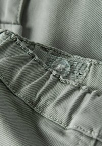 Grijze stof met een fijne textuur, voorzien van een duidelijke knoop en zichtbare stikdetails. De stof heeft een lichte glans.