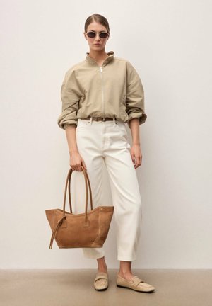 Mujer con chaqueta beige con cremallera, pantalones blancos, mocasines y gafas de sol, sosteniendo un gran bolso tote de cuero marrón, de pie frente a un fondo liso.