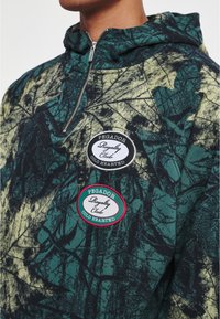 Sudadera con capucha en verde oscuro y amarillo con un patrón de hojas. Presenta dos parches de tela circulares con el texto "PEgador Royalty Club".
