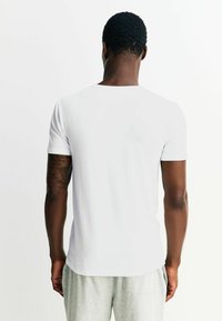 Balts īsroku T-krekls no mīksta kokvilnas materiāla, ar apaļu kaklu un gludu tekstūru, redzams no aizmugures, kopā ar pelēkām biksēm.