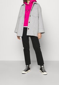 Šedý vlněný kabát s velkými kapsami, růžová mikina s kapucí a logem, černé kalhoty a černé vysoké tenisky s bílou podrážkou.