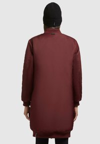 Bordeaux lang jack met een gladde, glanzende afwerking. Voorzien van ribgebreide manchetten en zoom. Draagt een zwarte gebreide beanie, gezien vanuit de rug.