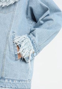 Giacca di denim azzurro chiaro con polsini sfrangiati, colletto classico, tasche frontali e una vestibilità comoda. Il tessuto appare morbido e testurizzato.