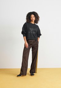 Hanorac negru cu un logo lucios, asortat cu pantaloni maro cu dungi galbene pe laterale și adidași negri, pe un fundal de podea galben.