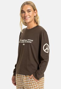 Camisetta marrone a maniche lunghe con scritte bianche e simbolo della pace sulla manica destra; materiale in cotone morbido; vestibilità comoda.