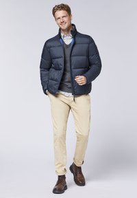 Marineblaue Daunenjacke mit hohem Kragen, Reißverschluss und gestepptem Material. Getragen über einem grauen Pullover und einem Hemd mit Kragen, kombiniert mit beigen Hosen und braunen Stiefeln.