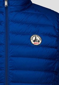 Chaqueta azul acolchada con un parche redondo que muestra a una persona en un scooter y el texto "Just over The Top JOTT" en el pecho.