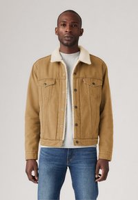 Chaqueta de mezclilla marrón con cuello de borrego, frontales abotonados, dos bolsillos en el pecho y un corte recto; llevada sobre una camisa gris y unos jeans azules.