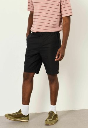 Person trägt schwarze Shorts, ein rosa gestreiftes kurzärmeliges Hemd, weiße Socken und olivgrüne Turnschuhe und steht mit den Händen in der Nähe der Taschen.