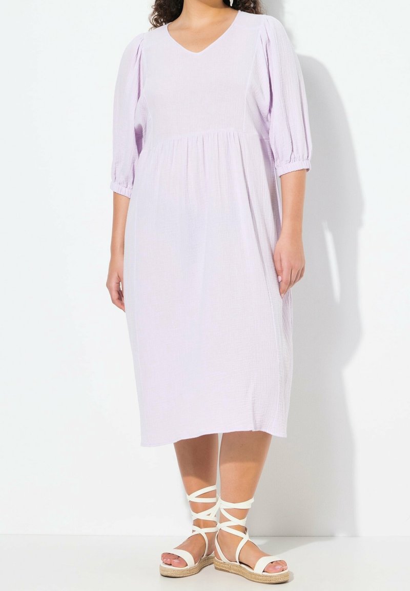 Lavendelfarbenes knielanges Kleid mit V-Ausschnitt, Puffärmeln und gerafftem Bund. Kombiniert mit weißen Sandalen mit Riemchen und einer natürlichen Jutesohle.