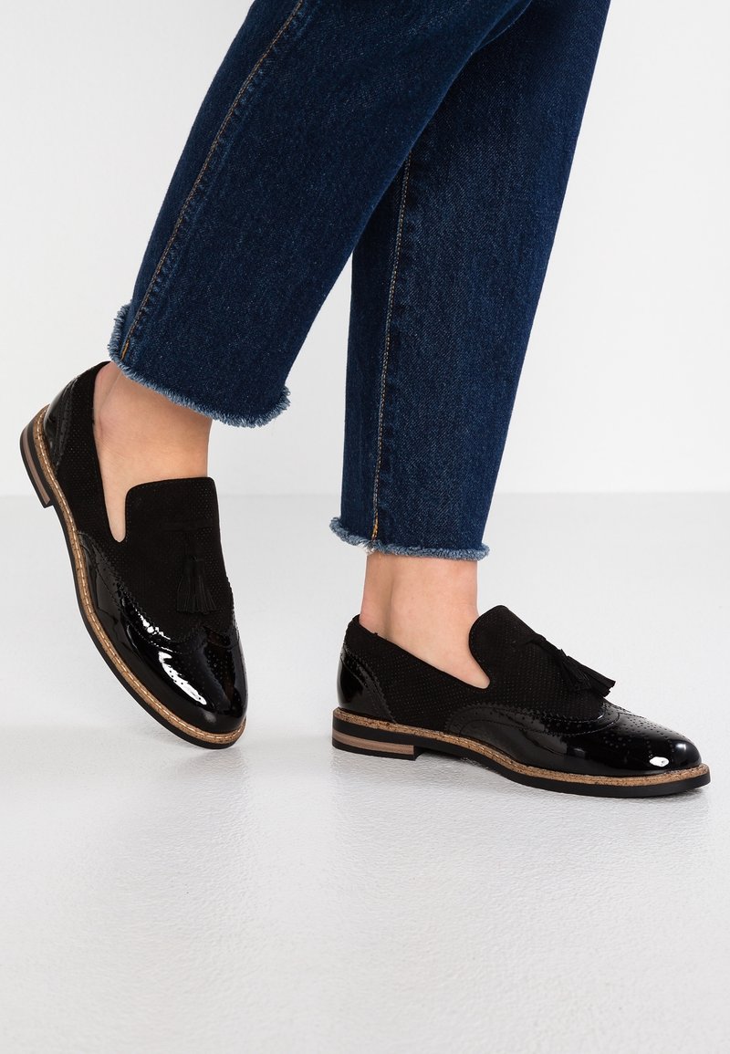 Jambes en jean bleu foncé raccourci portant des mocassins en cuir verni noir avec des glands et une semelle en bois, debout sur un sol clair.