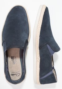 Chaussons en suède bleu marine avec un accent en jute tissé le long de la semelle et des panneaux latéraux élastiques pour un enfilage facile. Doublure intérieure beige et rembourrage.