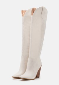 Bílé vysoké kozačky ke kolenům s špičatou špičkou, hladkým koženým materiálem a texturovaným, na sebe navazujícím klínovým podpatkem. Vrchní část je zdobena vroubkovaným detailem.
