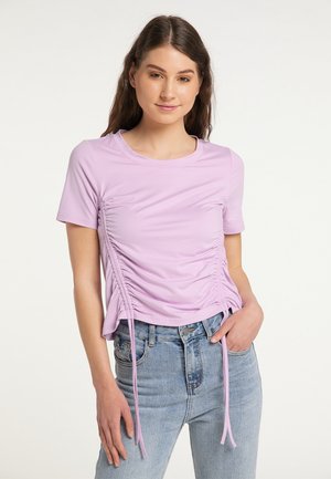 Junge Frau mit langen braunen Haaren trägt ein helllila gerafftes Kurzarmtop und hellblaue Jeans, posiert mit einer Hand in der Tasche.