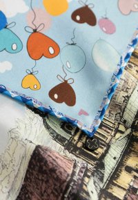 Twee gelaagde stoffen: de ene toont kleurrijke ballonnen en harten op blauw, de andere heeft een textuur van een sepia-tonig architectonisch ontwerp. Blauwe scalloped rand.