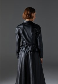 Čierny kožený outfit, ktorý zahŕňa priliehavú bundu s peplum pásom a dlhú, splývavú sukňu. Hladká textúra a elegantný dizajn.