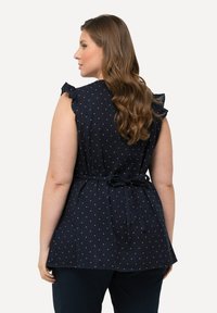 Blusa blu navy con pois bianchi, maniche corte arricciate, vita aderente e un fiocco annodato nella parte posteriore. Tessuto leggero con una finitura testurizzata.