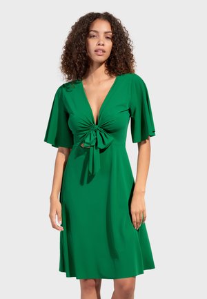 Kvinde med krøllet hår iført en grøn knælængde kjole med flagrende ærmer og en knude foran under halsudskæringen.