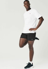 Hvid t-shirt med korte ærmer og et rødt logo, parret med sorte atletikshorts. Modellen bærer sorte sneakers med hvide såler.