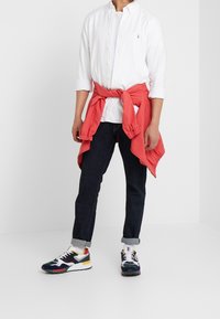 Witte overhemd met knopen, koraalkleurige trui om de taille gebonden, donkergroene spijkerbroek en meerkleurige sneakers met een gestructureerd ontwerp.