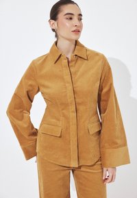 Ženska, oblečena v gorčično rumen kordurov suknjič in hlače, gleda v stran z zavezano pričesko na belem ozadju.