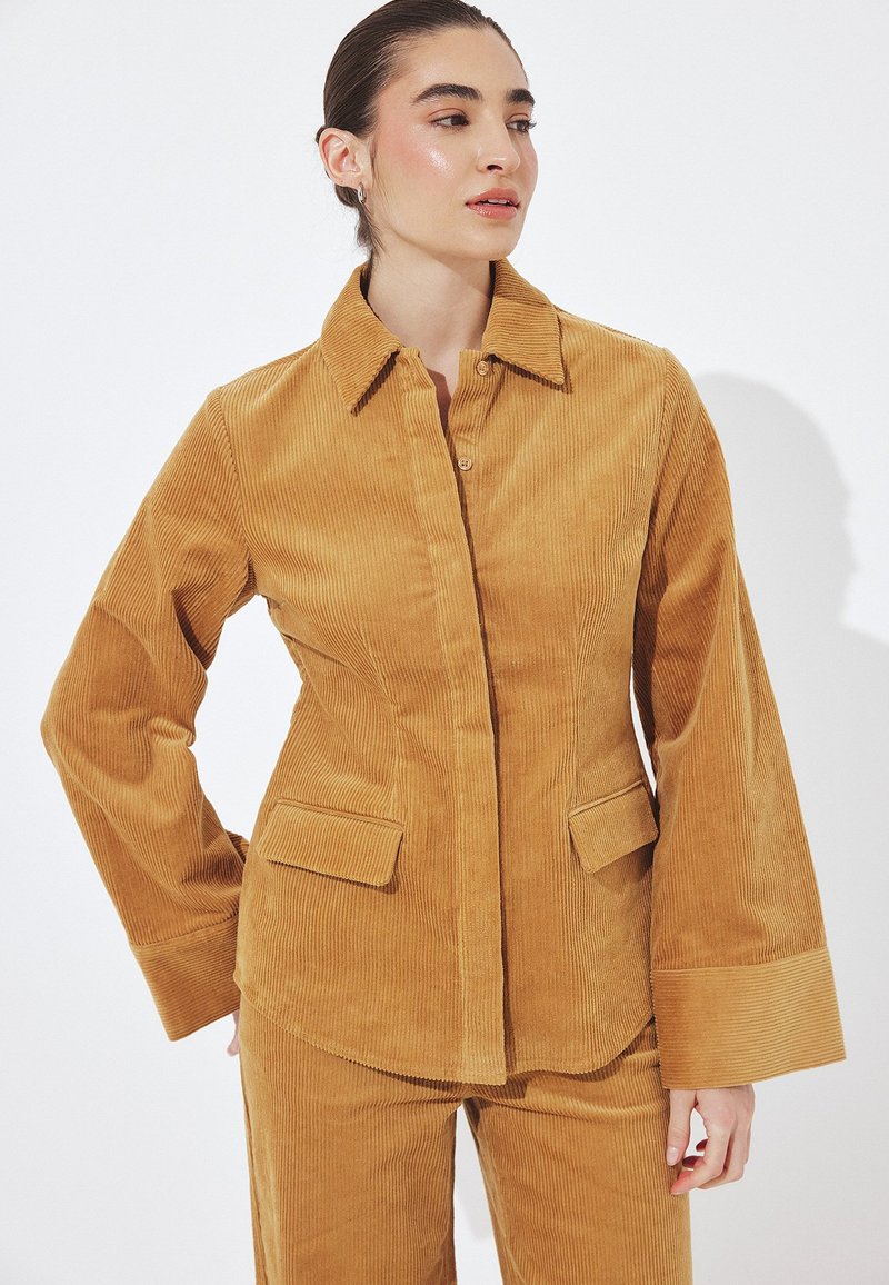 Ženska, oblečena v gorčično rumen kordurov suknjič in hlače, gleda v stran z zavezano pričesko na belem ozadju.