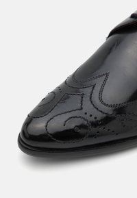 Scarpa di pelle nera con punta affilata, caratterizzata da intricate decorazioni impresse, perforazioni e cuciture in rilievo lungo i bordi.