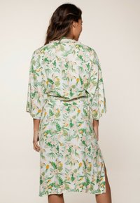 Kimono de flores con patrones tropicales en verde y amarillo sobre un fondo claro, con mangas anchas y cinturón en la cintura, diseño hasta la rodilla.