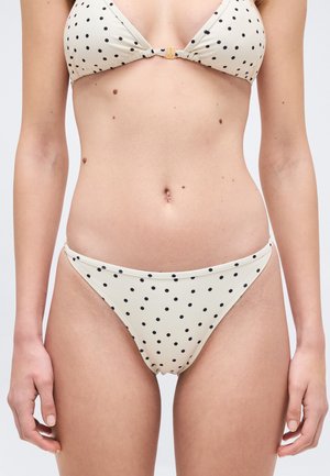 Γυναίκα που φοράει λευκό μπικίνι με μαύρες πουά, δείχνοντας τον κορμό και το πάνω μέρος των ποδιών της μπροστά σε απλό φόντο.