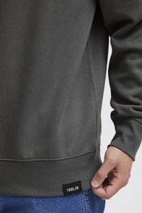 Gray sweatshirt with ribbed cuffs and hem, side panel details, and a black brand tag at the lower left. Visible hand gripping the hem.