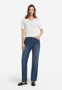 Hvid kortærmet V-hals T-shirt, parret med lige ben mørke denim jeans og sorte flettede sandaler, stående på en ensfarvet baggrund.