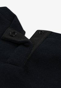 Juodas megztas audinys su struktūrizuotu apykaklės dizainu, kuriame yra du juodi spaustukai prie iškirptės. Audinio tekstūra yra švelni ir vienoda visame paviršiuje.