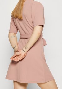 Krátké rukávy, upnuté šaty z měkké růžové látky s rozšířenou sukní, zúženým pasem a uvázaným páskem. Obsahují viditelné tetování na rukou.