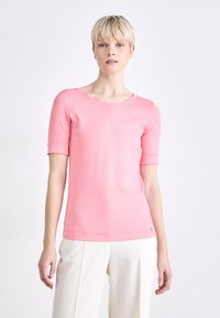 Roze t-shirt met korte mouwen en een ronde hals, gladde structuur en een logo detail op de onderkant links. Gecombineerd met witte brede broek.