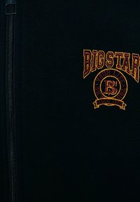 Svart zip-jacka med ribbat mönster, broderad "BIG STAR"-logotyp i gult och vinrött, med detaljer om året "1979" nedanför.