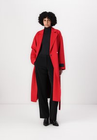 Roter Wollmantel mit einem langen, lockeren Schnitt, Reverskragen und einem einzelnen Knopfdetails, kombiniert mit schwarzen Hosen und schwarzen Schuhen.