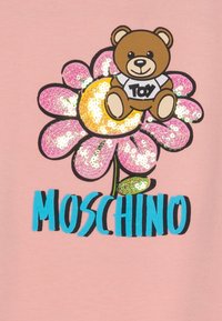 Růžový oděv s velkým designem květiny pokryté flitry, s kresleným medvědem držícím štítek označený "Hračka" a silným modrým textem "MOSCHINO".