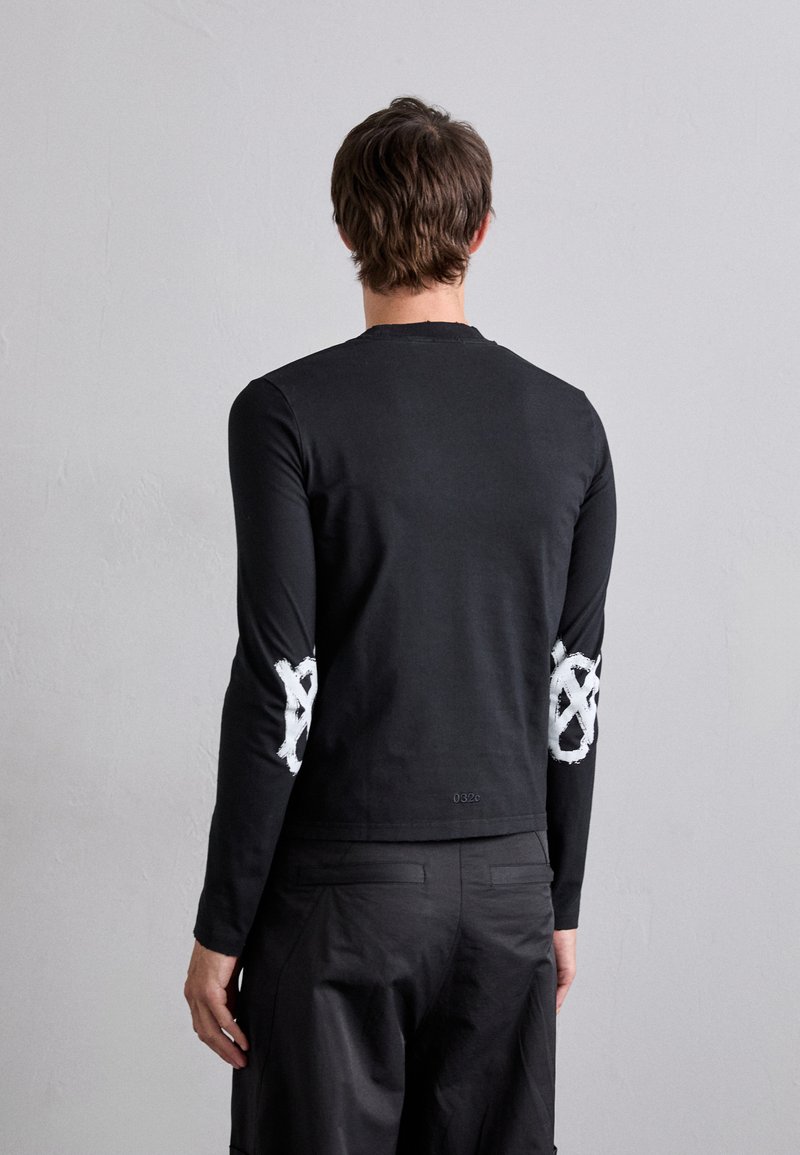 Schwarzes Langarmshirt aus Baumwolle. Hat weiße, abstrakte Designs auf den Ellbogen. Minimalistisches Design mit Rundhalsausschnitt und schmaler Passform.