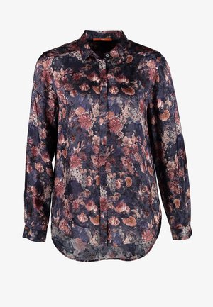 Persona con blusa oscura de flores, vaqueros ajustados azules y botines marrones, sosteniendo un sombrero marrón, de pie contra un fondo liso.