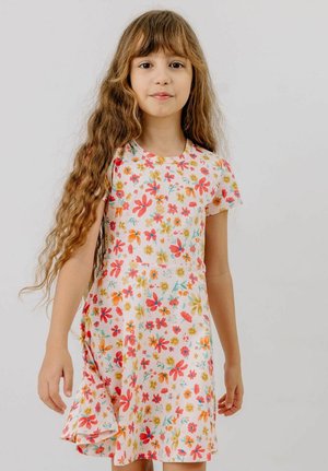 Young girl with long wavy hair wearing a white dress covered in colorful floral patterns, standing against a plain background.