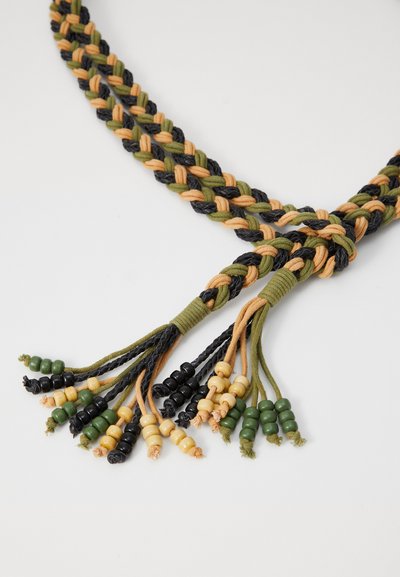 Accessoire textile tressé en noir, vert et beige avec plusieurs pompons. Comprend des perles en bois de couleur verte, noire et marron clair.
