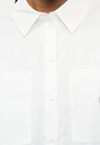 Nærbillede af en person iført en skarp hvid skjorte med knapper, spids krave og to forlommer.