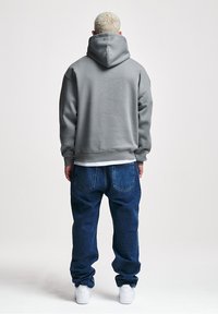 Felpa grigia con cappuccio realizzata in un materiale morbido, abbinata a jeans blu larghi e sneakers bianche, vista da dietro.