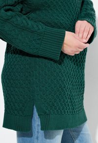 Maglione verde a trecce con un motivo texturizzato, polsini e orlo a coste, con spacchi sui lati. Indossato sopra jeans in denim blu.