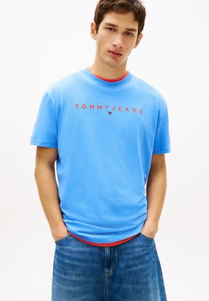 Fiatal férfi világoskék Tommy Jeans pólóban piros felirattal, kék farmerben, kezét zsebében tartva, fehér háttér előtt.