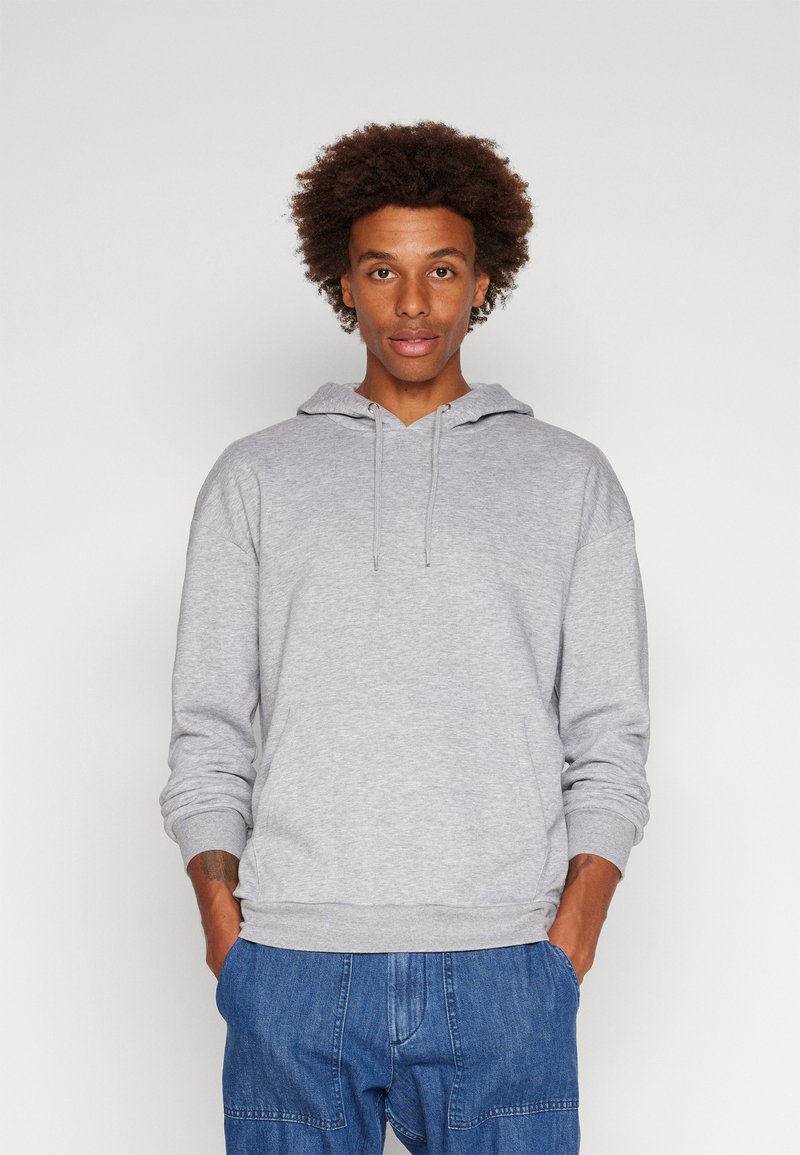 Felpa grigia chiara con cappuccio regolabile, polsini e orlo a costine, realizzata in un tessuto morbido, caratterizzata da una vestibilità rilassata. Abbinata a pantaloni in denim blu.