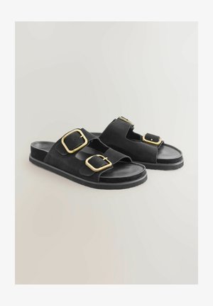 Sandales en suedine noire avec deux larges courroies et boucles en or, une semelle plate et un design ouvert au niveau des orteils. Texture lisse avec des accents de quincaillerie contrastants.