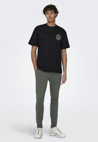 Camiseta negra de algodón con un estampado gráfico verde en el lado izquierdo, combinada con pantalones ajustados verde claro y zapatillas deportivas blancas.